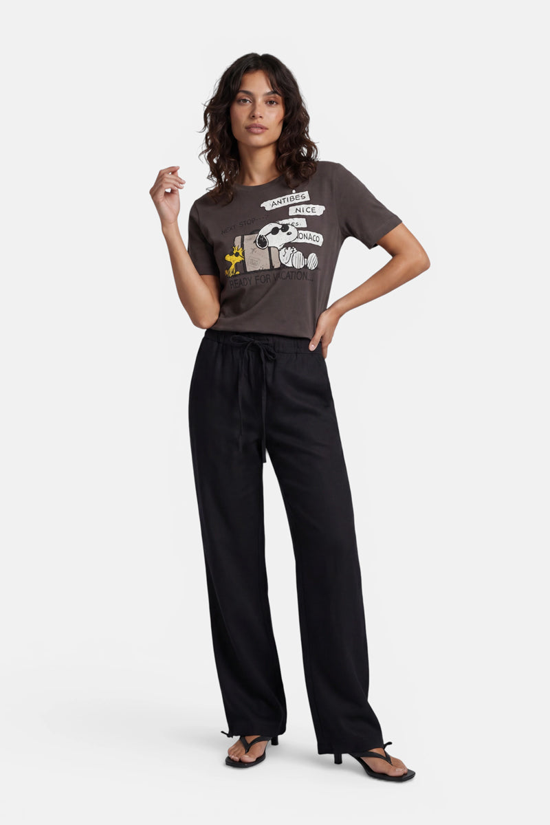 Eine Frau mit welligem, dunklem Haar posiert vor einem weißen Hintergrund in einem limitierten Snoopy-T-Shirt aus Baumwolle von Princess Goes Hollywood, dazu trägt sie eine weite schwarze Hose und schwarze Sandalen; eine Hand hat sie im Haar, die andere auf der Hüfte.