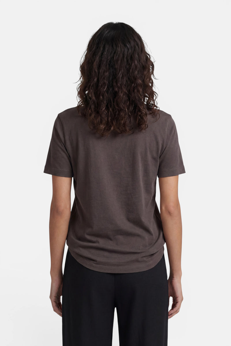 Eine Person mit schulterlangen, lockigen braunen Haaren steht mit dem Rücken zur Kamera und trägt ein dunkelbraunes Baumwoll-T-Shirt mit kurzen Ärmeln aus der limitierten Auflage „Princess Goes Hollywood“ sowie eine schwarze Hose. Vor einem weißen Hintergrund hängen ihre Arme entspannt an den Seiten herab.