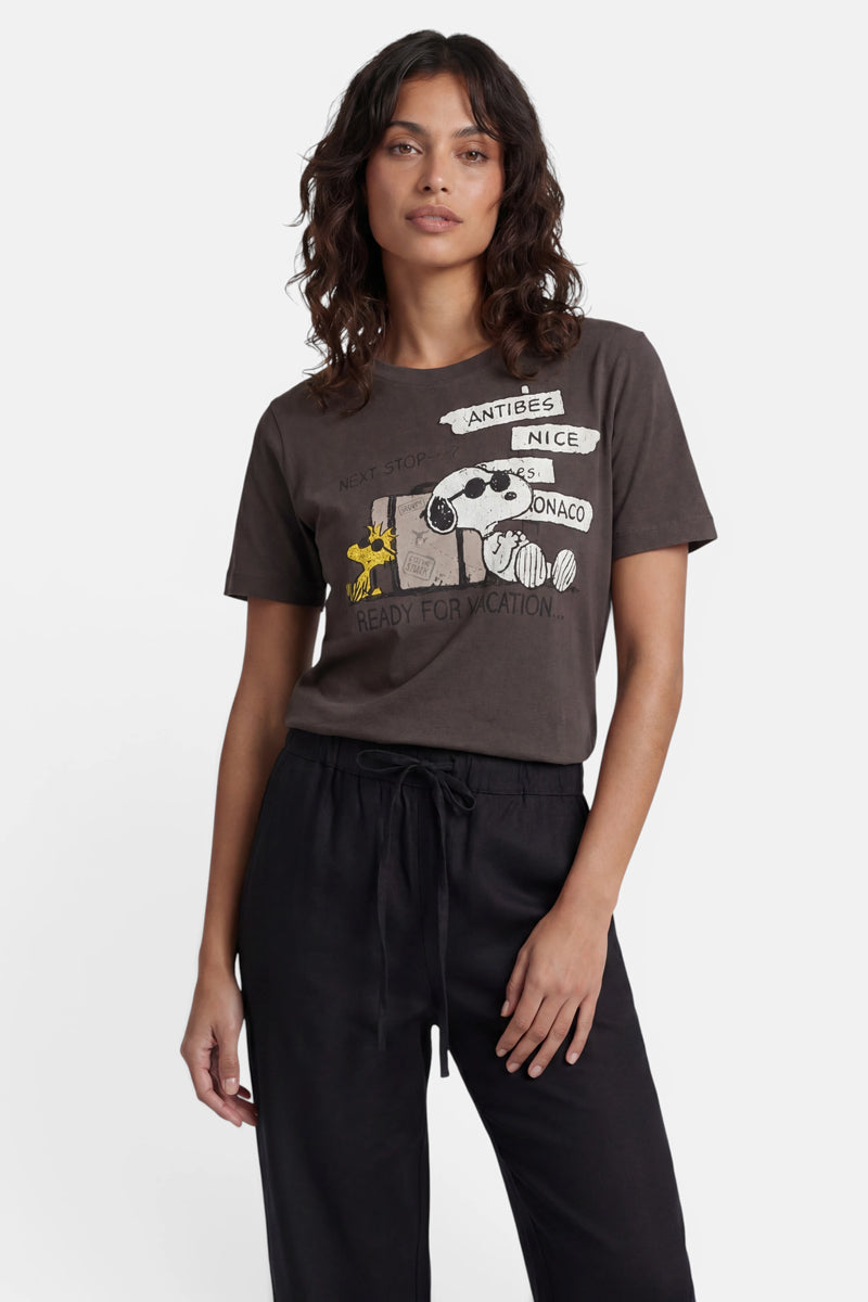 Eine Frau mit welligem, dunklem Haar trägt ein „Princess Goes Hollywood Limited Edition“-Snoopy-T-Shirt aus Baumwolle, auf dem Snoopy und Woodstock mit Koffern zu sehen sind, mit der Aufschrift: „Nächster Halt: Antibes, Nizza, Monaco. Bereit für den Urlaub.“ Dazu trägt sie eine schwarze Hose vor einem weißen Hintergrund.