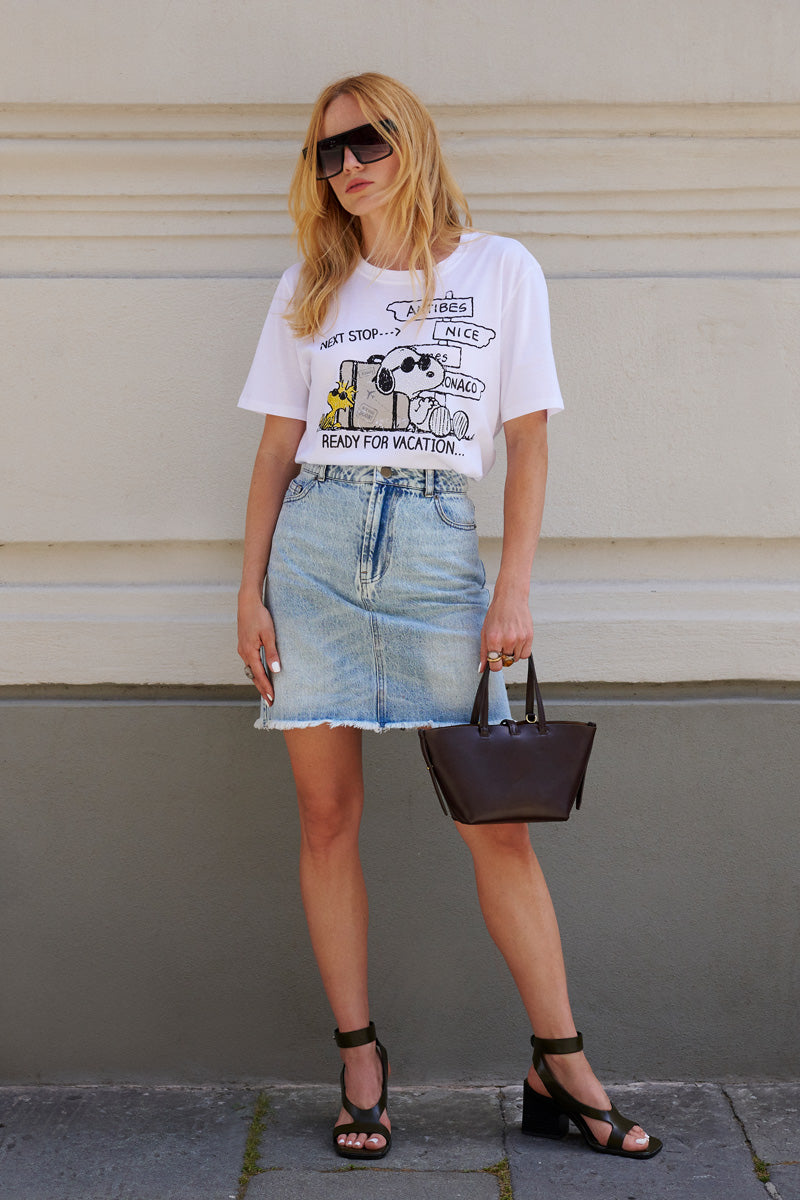 Eine Frau mit langen blonden Haaren und einer großen Sonnenbrille lehnt entspannt an einer hellbeigen Wand. Sie trägt ein „Princess Goes Hollywood“-Snoopy-T-Shirt in limitierter Auflage, einen hellblauen Jeansrock und schwarze Riemchensandaletten und hält eine dunkelbraune Handtasche in der Hand.