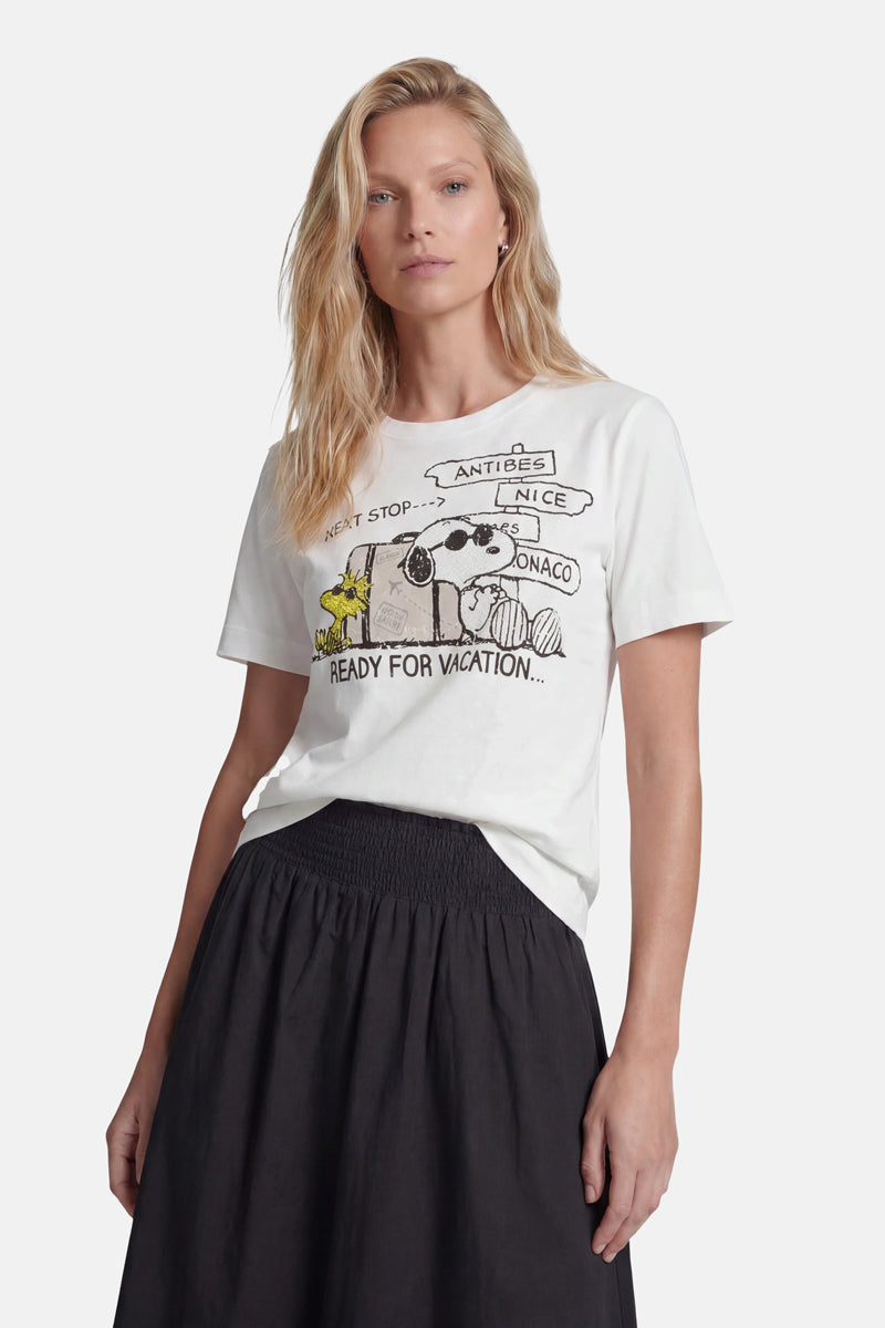 Eine Frau mit langen blonden Haaren trägt ein weißes Snoopy-T-Shirt aus der „Princess Goes Hollywood“-Limited-Edition, auf dem Cartoon-Tiere in einem Auto zu sehen sind und der Schriftzug „Nächster Halt … Antibes, Nizza, Monaco“ steht. Dazu trägt sie einen schwarzen Rock mit hoher Taille vor einem weißen Hintergrund.