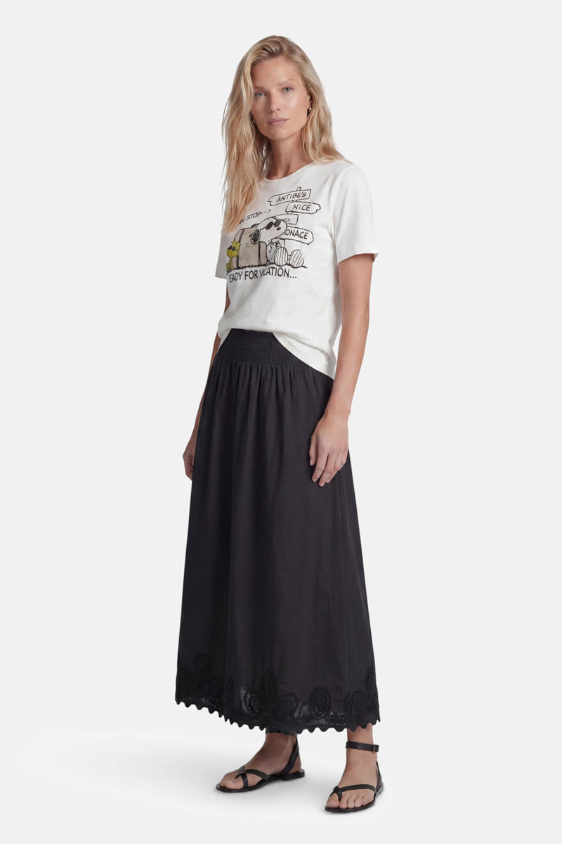 Eine Frau mit langen blonden Haaren blickt nach vorne. Sie trägt ein „Princess Goes Hollywood Limited Edition Snoopy“-T-Shirt aus Baumwolle und einen schwarzen, hoch taillierten, knöchellangen Rock mit Wellenrand, dazu schwarze Sandalen – vor einem schlichten weißen Hintergrund.