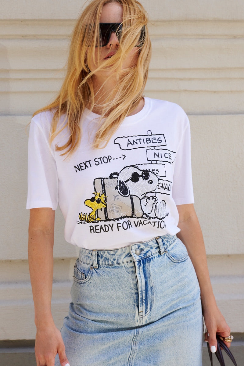 Eine Frau mit langen blonden Haaren, die ihr Gesicht teilweise verdecken, trägt eine schwarze Sonnenbrille und ein „Princess Goes Hollywood Limited Edition Snoopy“-Baumwoll-T-Shirt mit Woodstock, Koffern und französischen Verkehrsschildern, dazu einen hellblauen Jeansrock vor einer hellen Wand.