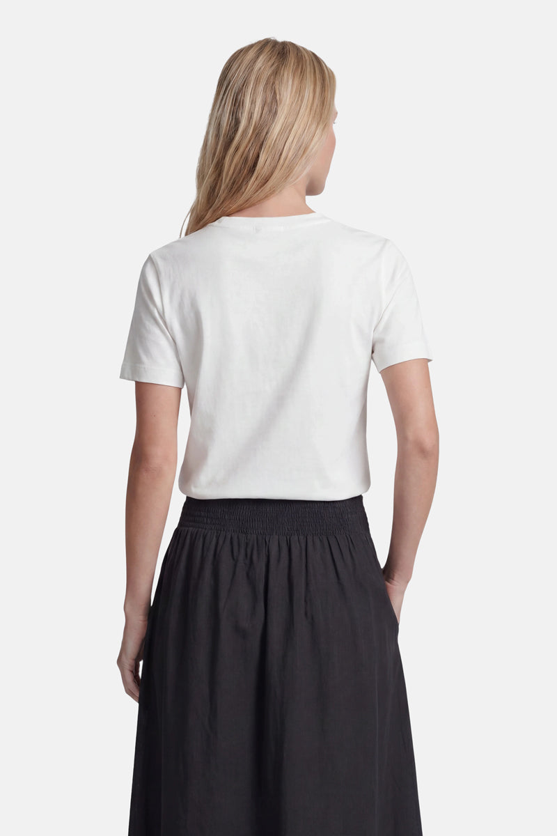 Vor einem weißen Hintergrund ist eine Person mit langen, hellbraunen Haaren von hinten zu sehen, die ein schlichtes weißes „Princess Goes Hollywood“-Baumwoll-Snoopy-T-Shirt trägt, das in einen schwarzen Rock mit hoher Taille und gerafftem Bund gesteckt ist.