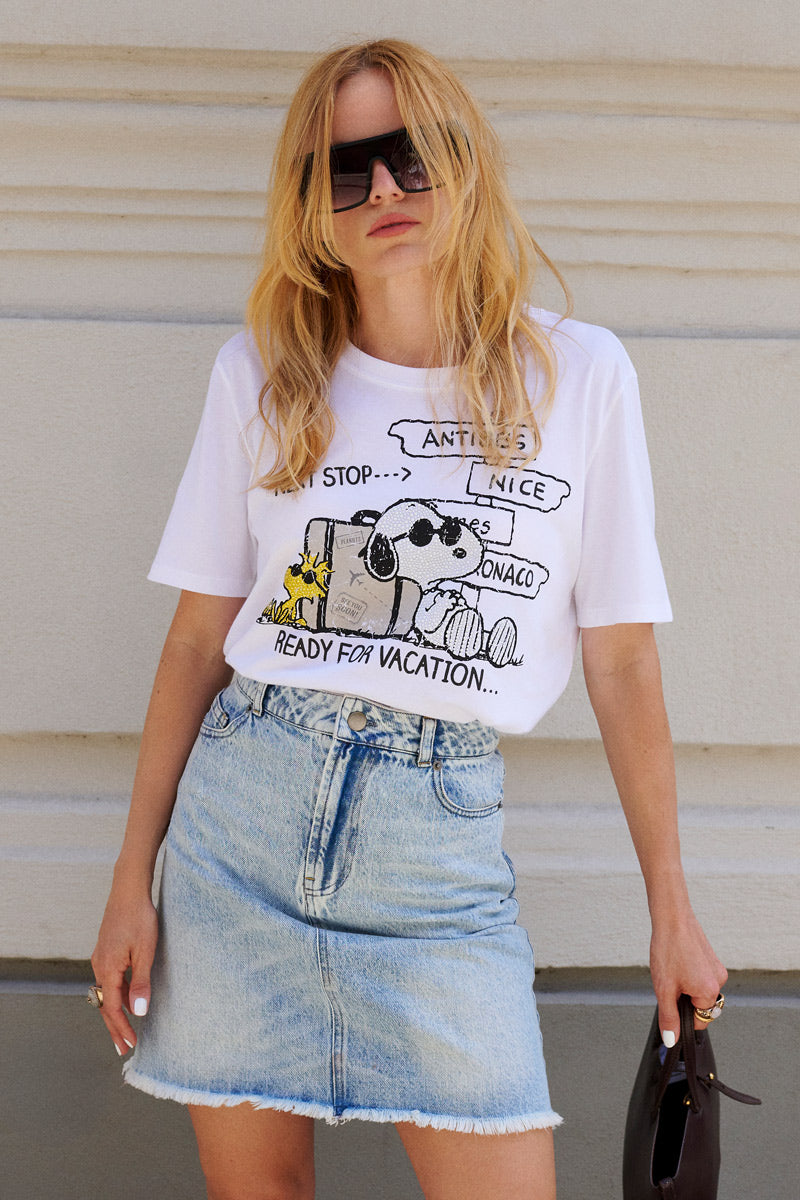 Eine Frau mit langen blonden Haaren und einer großen Sonnenbrille trägt ein „Princess Goes Hollywood“-Snoopy-T-Shirt aus Baumwolle in limitierter Auflage, dazu einen hellblauen Jeansrock mit hoher Taille. Sie steht entspannt vor einer hellen Wand und hält eine kleine Tasche in der Hand.