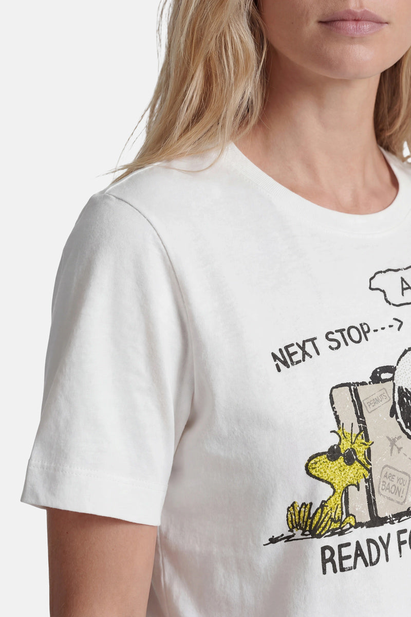Eine hellhäutige, blonde Frau trägt ein Snoopy-T-Shirt aus Baumwolle der Limited Edition „Princess Goes Hollywood“, auf dem Woodstock neben einem Koffer abgebildet ist. Auf dem Shirt steht „NEXT STOP... READY F“, wobei Teile der Grafik und des Textes abgeschnitten sind; nur ihr unterer Gesichtsbereich ist zu sehen.