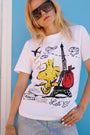 Eine Frau mit übergroßer Sonnenbrille und heller Jeans trägt ein T-Shirt aus der „Princess Goes Hollywood“-Limited-Edition mit Woodstock-Print und Pailletten, auf dem „Let’s Go!“ steht, während sie am Eiffelturm entlangschlendert und dabei ein rotes Bündel in der Hand hält – ganz in der Nähe einer leuchtend blauen Wand.