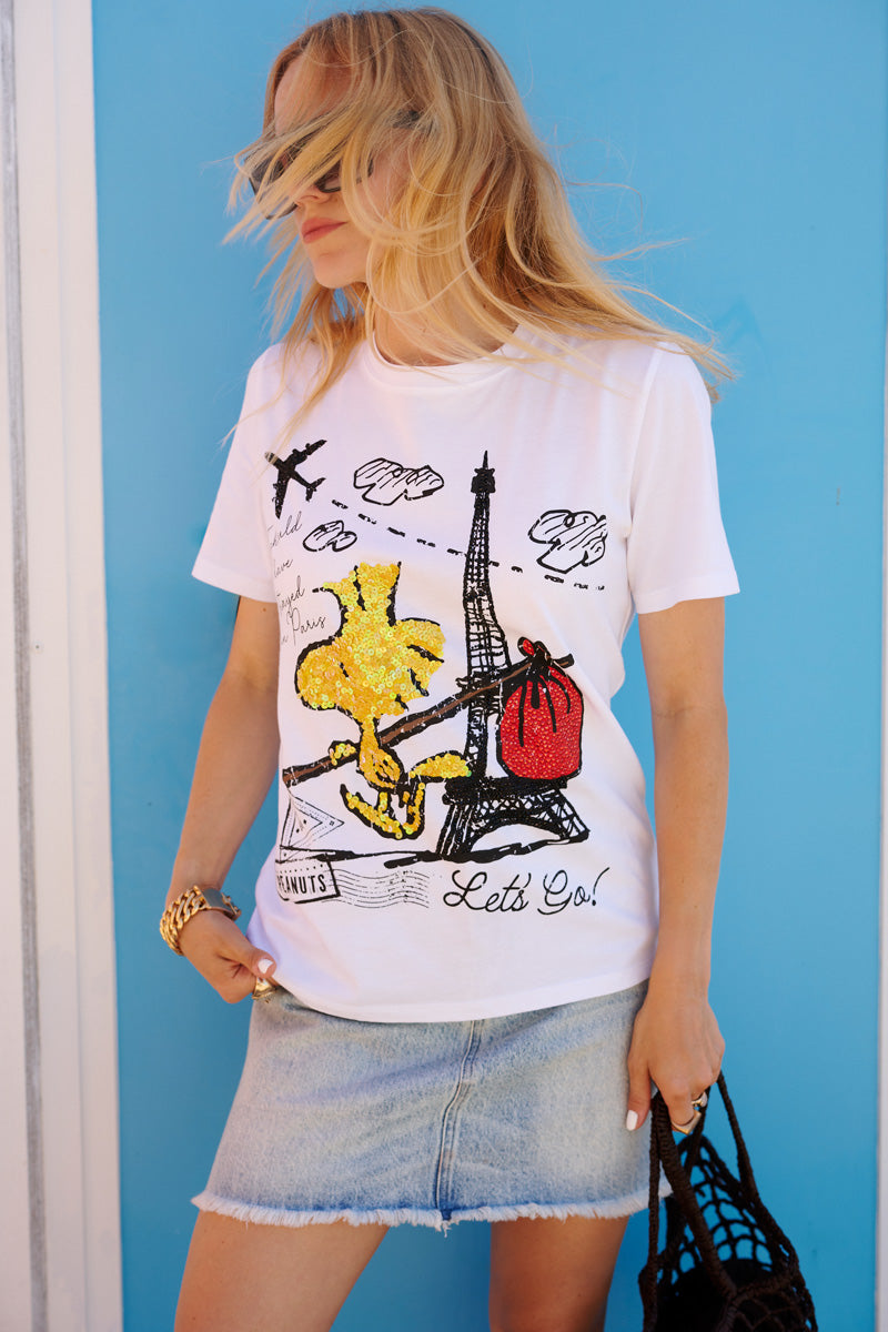 Eine blonde Frau steht vor einer strahlend blauen Wand und trägt ein weißes T-Shirt der Limited Edition „Princess Goes Hollywood“ mit einem Aufdruck des Eiffelturms und von Woodstock, dazu einen hellen Jeans-Minirock und eine schwarze Flecht-Tasche.