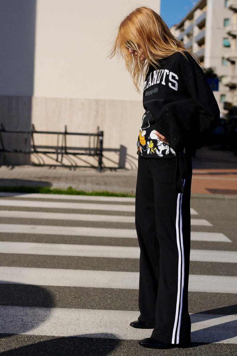 Eine Person mit langen blonden Haaren steht auf einem Zebrastreifen und trägt ein schwarzes Sweatshirt der Limited Edition „Princess Goes Hollywood“ mit Comic-Motiven und dem Schriftzug „GIANTS“, eine schwarze Jogginghose mit weißen Streifen sowie schwarze Schuhe. Im Hintergrund sind Gebäude und ein Zaun zu sehen.