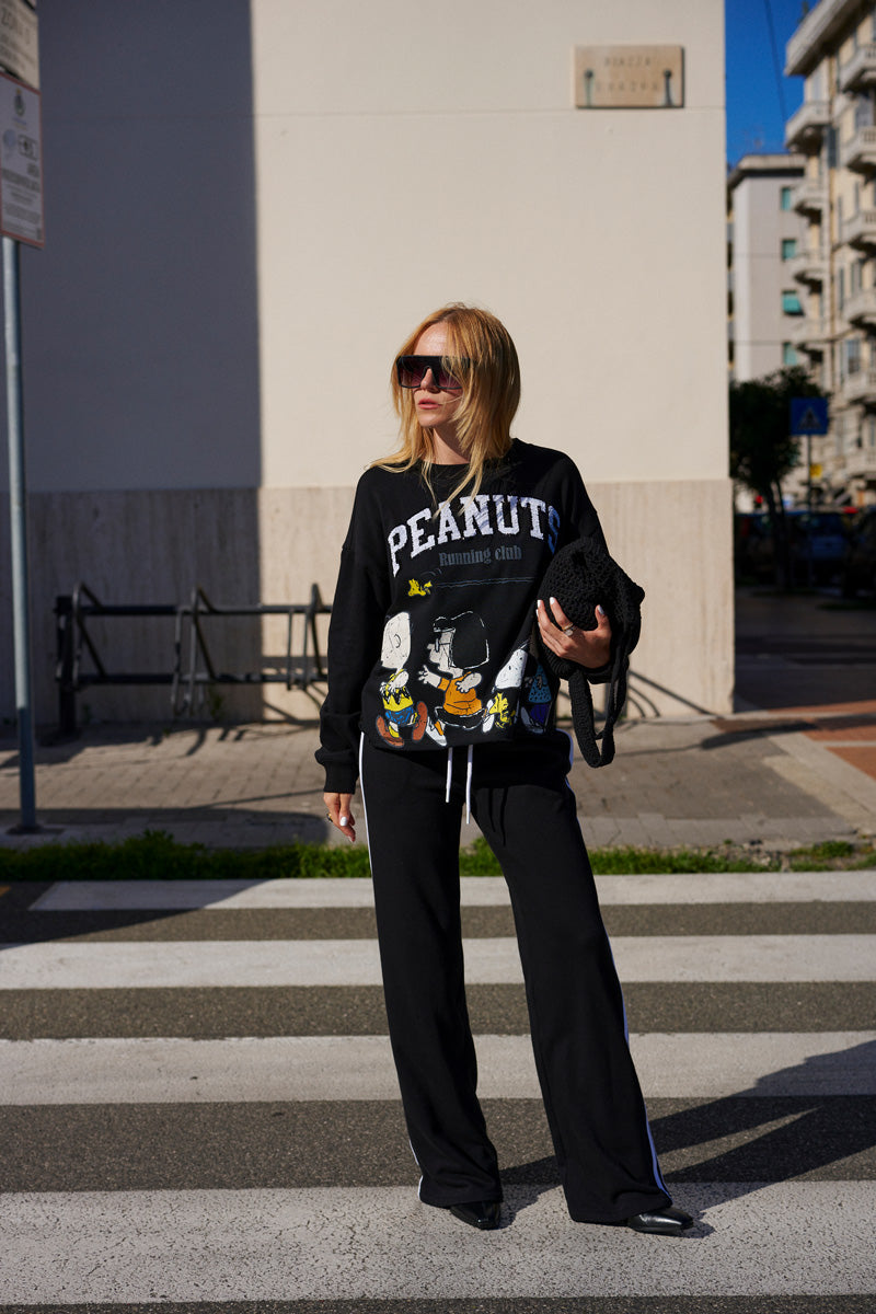 Eine Frau mit blonden Haaren und Sonnenbrille steht auf einem Zebrastreifen und trägt einen Pullover aus der „Princess Goes Hollywood Limited Edition Peanuts“-Kollektion – einen übergroßen Sweater mit Comicfiguren –, dazu eine schwarze Hose mit weitem Bein und eine schwarze, gesteppte Tasche.