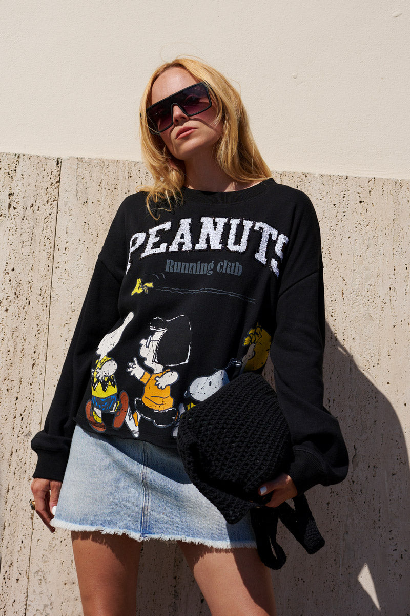Eine Frau mit einer übergroßen Sonnenbrille und einem schwarzen „Princess Goes Hollywood Limited Edition“-Peanuts-Pullover mit Comicfiguren, dazu einen hellen Jeans-Minirock, steht mit einer schwarzen, strukturierten Tasche in der Hand vor einer beigen Marmorwand. Das Sonnenlicht wirft Schatten hinter ihr.