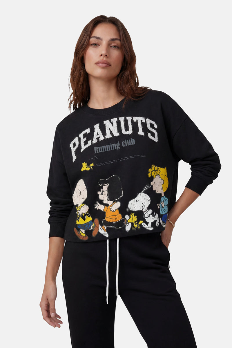 Eine Frau mit mittellangem braunem Haar trägt ein schwarzes Sweatshirt aus der „Princess Goes Hollywood“-Limited-Edition, auf dem über den „Peanuts“-Figuren der Schriftzug „PEANUTS Running Club“ zu sehen ist. Sie steht mit einer Hand in der Hosentasche vor einem schlichten Hintergrund.
