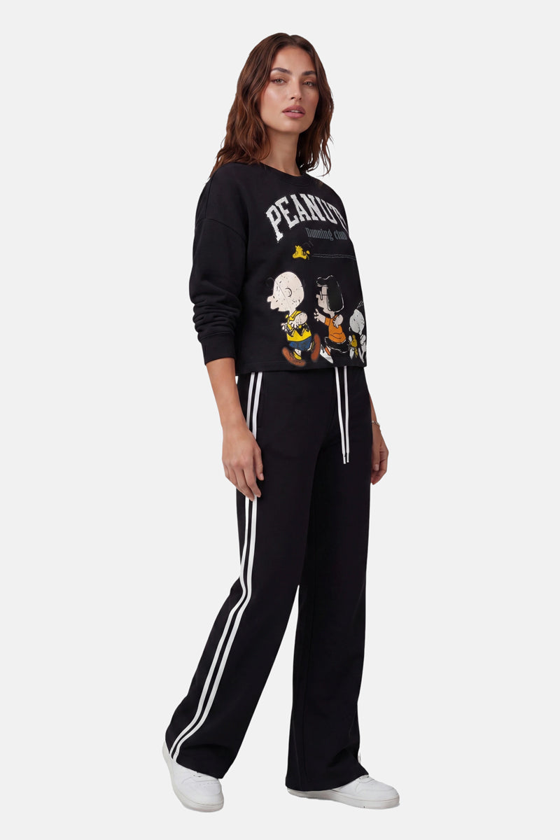 Eine Frau mit welligem braunem Haar und ausdruckslosem Gesicht trägt ein schwarzes „Peanuts“-Sweatshirt von Princess Goes Hollywood aus Baumwolle mit Comicfiguren, eine schwarze Jogginghose mit weißen Seitenstreifen und weiße Sneaker vor einem hellen Hintergrund.