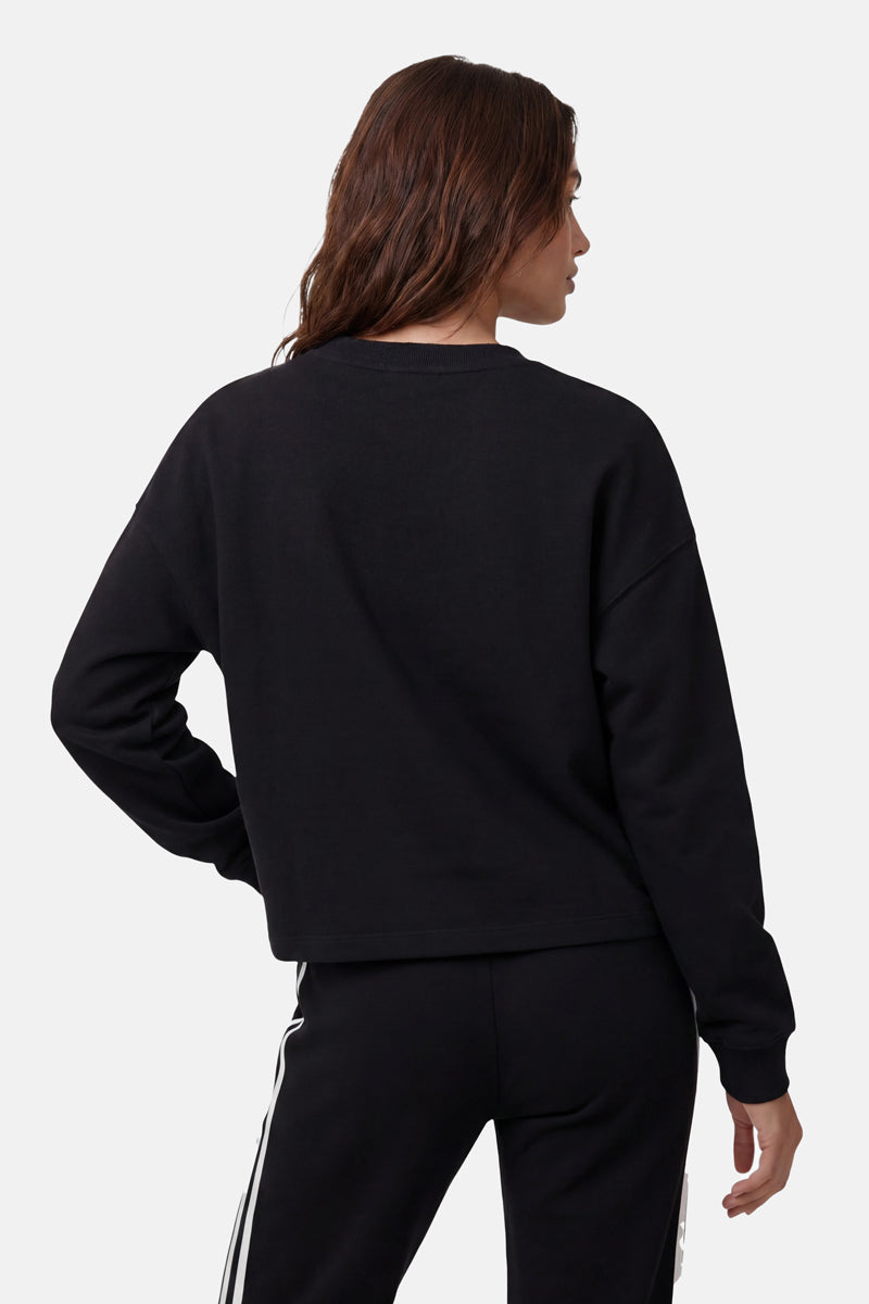 Eine Frau mit langen braunen Haaren, von hinten gesehen, trägt einen übergroßen Sweater aus der „Princess Goes Hollywood“-Limited-Edition und eine dazu passende schwarze Hose mit weißen Streifen. Sie steht vor einem schlichten hellgrauen Hintergrund und hat den Kopf leicht nach links gedreht.