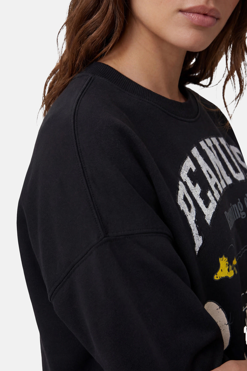 Eine Frau mit langen braunen Haaren trägt einen Pullover der „Princess Goes Hollywood“-Limited-Edition-Kollektion mit Peanuts-Motiven in verblasstem Schwarz mit weißer Schrift im Used-Look und teilweisen Comic-Motiven. Sie ist vor einem einfarbigen Hintergrund von den Schultern aufwärts zu sehen, die Lippen geschlossen.