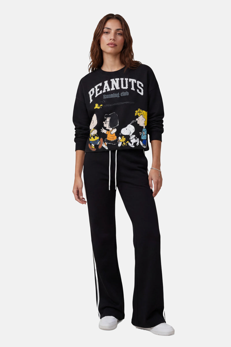 Eine Frau mit mittellangem braunem Haar und einem entspannten Gesichtsausdruck präsentiert vor weißem Hintergrund ein schwarzes, übergroßes Sweatshirt der „Princess Goes Hollywood Limited Edition PEANUTS Running Club“ mit Peanuts-Figuren, eine schwarze Hose mit weitem Bein und weißen Streifen sowie weiße Sneaker.