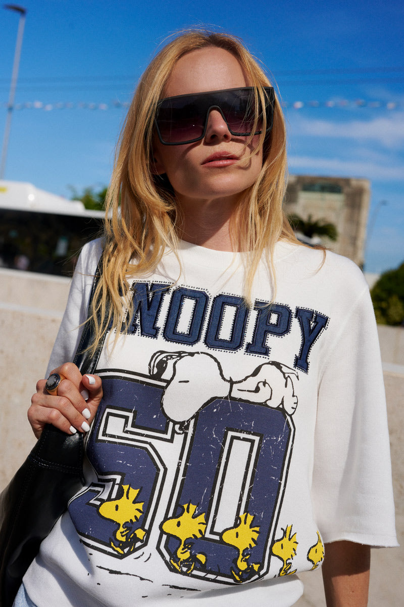 Eine blonde Frau trägt im Freien einen Snoopy-Sweater aus der Limited Edition „Princess Goes Hollywood“ – aus weißer Baumwolle mit Snoopy, einer blauen „50“ und Woodstock-Vögeln – sowie eine große schwarze Sonnenbrille und trägt eine schwarze Tasche unter einem strahlend blauen Himmel.
