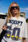 Eine blonde Frau trägt im Freien einen Snoopy-Sweater aus der Limited Edition „Princess Goes Hollywood“ – aus weißer Baumwolle mit Snoopy, einer blauen „50“ und Woodstock-Vögeln – sowie eine große schwarze Sonnenbrille und trägt eine schwarze Tasche unter einem strahlend blauen Himmel.