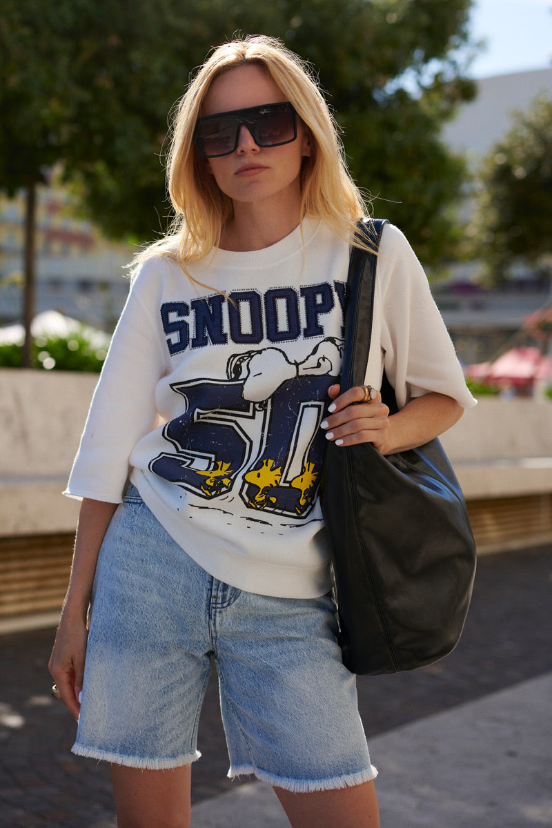 Eine Frau steht im Freien und trägt eine große schwarze Sonnenbrille, einen weißen Snoopy-Sweater aus Baumwolle aus der „Princess Goes Hollywood“-Limited-Edition, hellblaue Jeansshorts und eine große schwarze Umhängetasche. Im Hintergrund sind Bäume und eine verschwommene Stadtkulisse zu sehen.