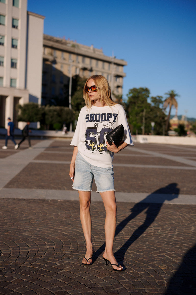 Eine Frau steht auf einem sonnigen Stadtplatz und trägt ein weißes „Princess Goes Hollywood Limited Edition“-Snoopy-50-T-Shirt aus Baumwolle, knielange Shorts aus hellem Denim, schwarze Sandalen mit Absatz und eine große Sonnenbrille. Sie hält eine schwarze Clutch in der Hand, hinter ihr sind Bäume und Türme zu sehen.