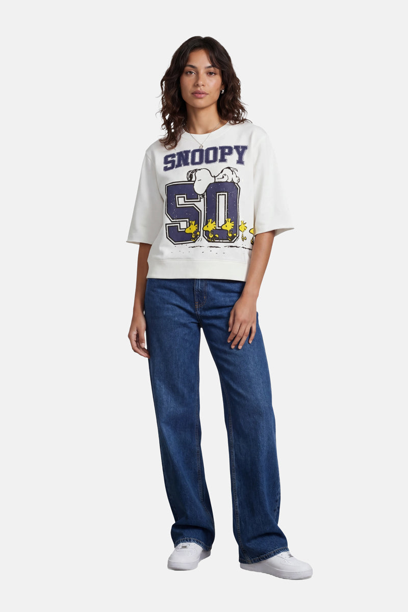 Eine Frau mit schulterlangem, gewelltem Haar und ausdruckslosem Gesicht präsentiert den weißen Snoopy-50-Sweater aus Baumwolle der Limited Edition „Princess Goes Hollywood“ mit einem Snoopy-Cartoon-Motiv, kombiniert mit blauen Jeans und weißen Sneakers.