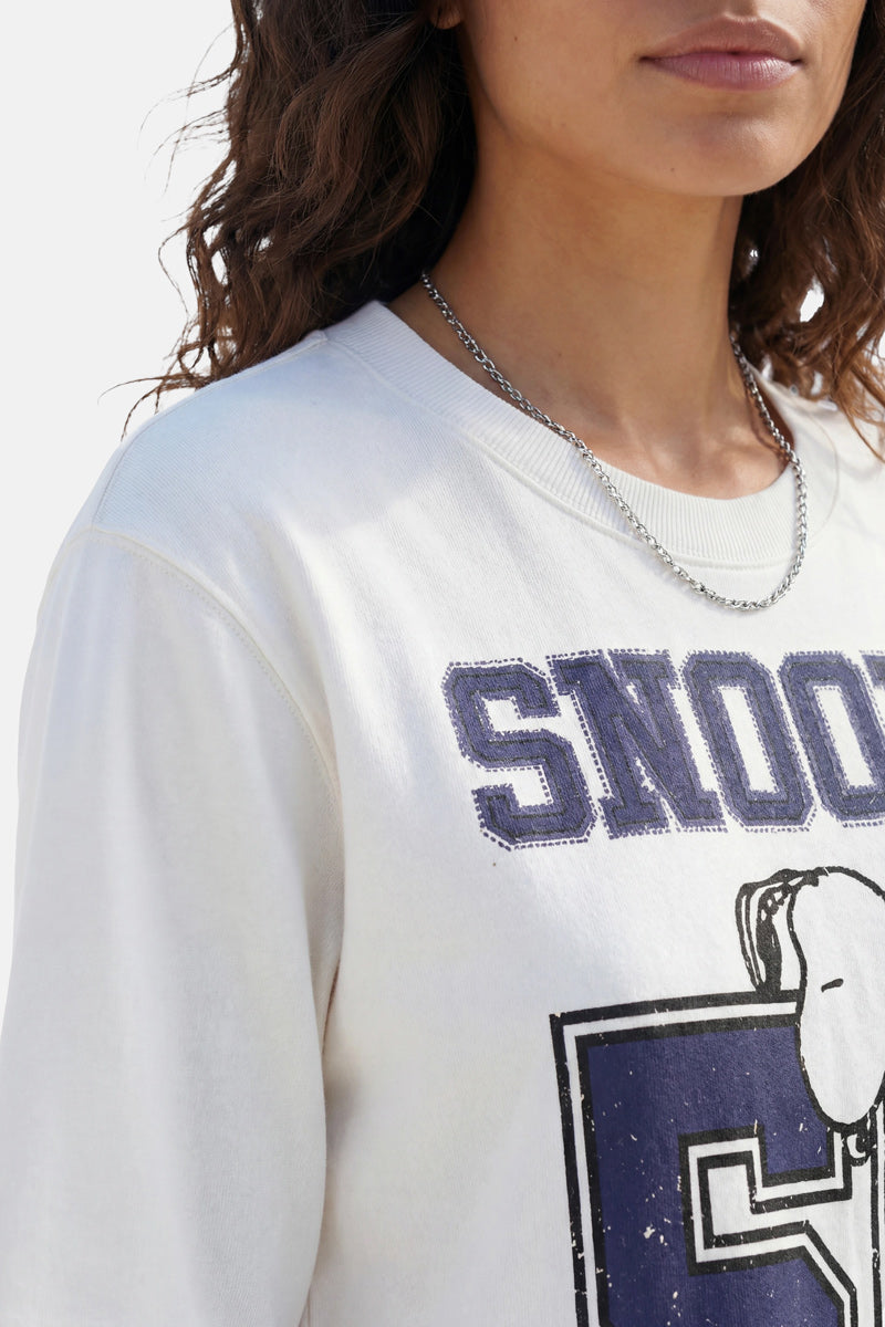 Eine Frau mit welligem braunem Haar trägt eine silberne Kette und einen Snoopy-Sweater aus Baumwolle aus der „Princess Goes Hollywood“-Limited-Edition mit auffälligem „SNOOP“-Schriftzug und einer Teilansicht der Snoopy-Figur. Das Bild zeigt ihre Lippen, ihren Hals und ihre Schultern.