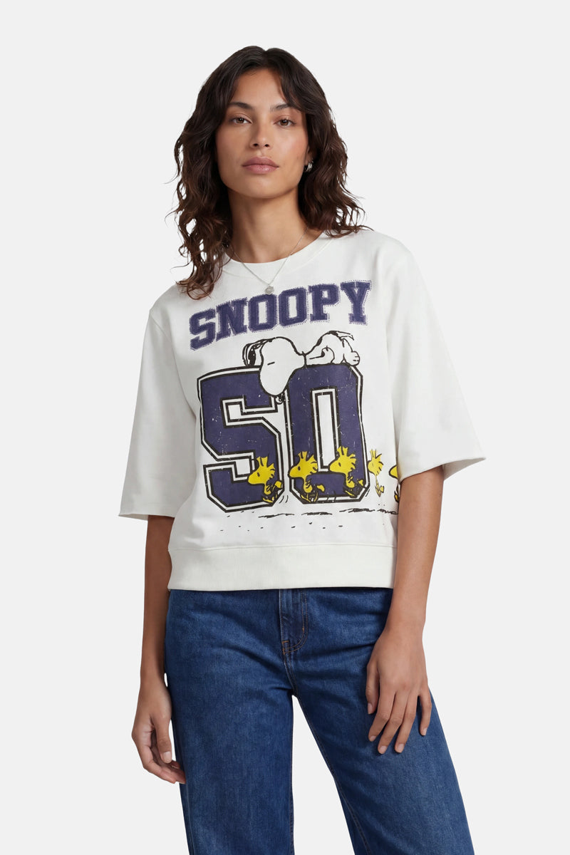 Eine Frau mit welligem, dunklem Haar trägt ein weißes T-Shirt der Limited Edition „Princess Goes Hollywood“, auf dem Snoopy auf einer großen 50 und darunter vier Woodstocks abgebildet sind. Auf der Brust prangt in fetten Buchstaben „SNOOPY“. Sie kombiniert dieses Baumwoll-T-Shirt mit einer blauen Jeans.