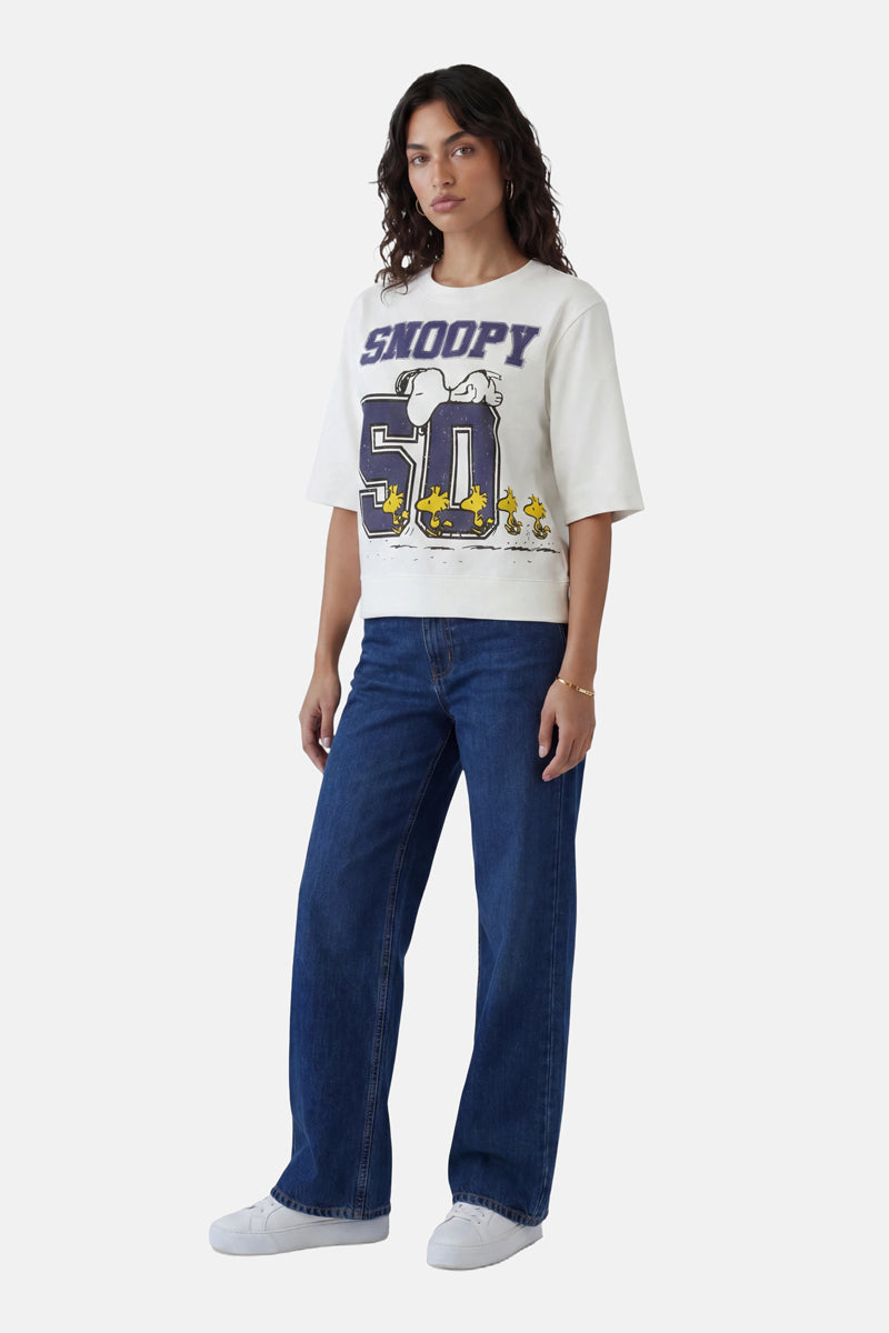 Vor einem schlichten weißen Hintergrund steht eine Frau mit gewelltem Haar und nur wenig Schmuck, die nach vorne blickt. Sie trägt einen Snoopy-Sweater aus Baumwolle aus der „Princess Goes Hollywood“-Limited-Edition, dunkelblaue Jeans und weiße Sneaker.