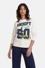 Eine Frau mit welligem braunem Haar trägt selbstbewusst einen Snoopy-Sweater aus Baumwolle aus der „Princess Goes Hollywood“-Limited-Edition, auf dem Snoopy auf einer großen „50“ zu sehen ist, darunter Woodstock und darüber „SNOOPY“ in Blau, dazu eine marineblaue Hose.