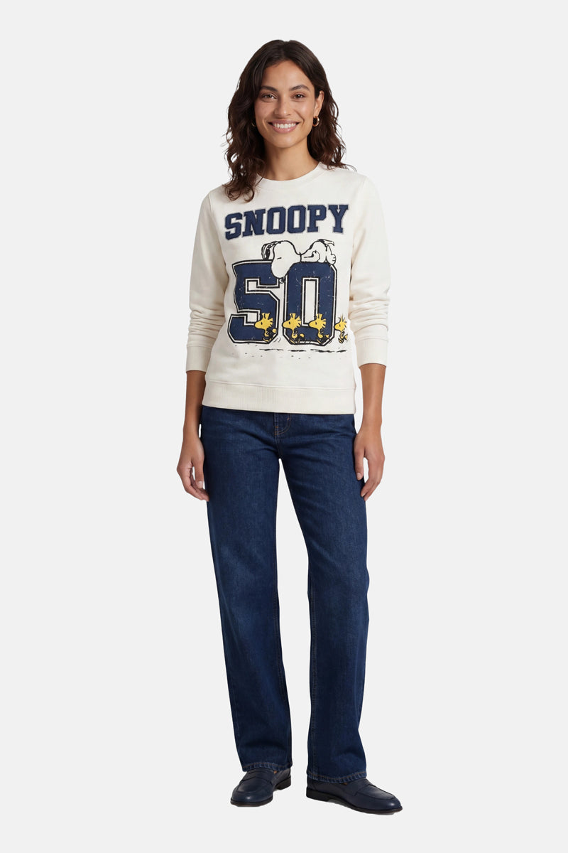 Eine Frau mit welligem braunem Haar lächelt in einem Snoopy-Sweatshirt aus Baumwolle der Limited Edition „Princess Goes Hollywood“, auf dem Woodstock und die Zahl 50 abgebildet sind. Dazu trägt sie eine dunkelblaue Jeans und schwarze Schuhe. Der Hintergrund ist schlicht weiß.