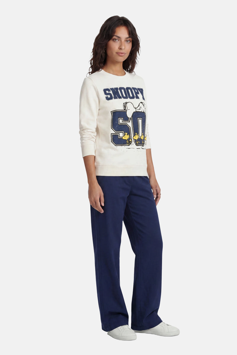 Vor einem schlichten weißen Hintergrund steht eine Frau mit welligem dunklem Haar und blickt nach vorne. Sie trägt einen Snoopy-Sweater aus Baumwolle aus der „Princess Goes Hollywood“-Limited-Edition mit „Snoopy 50“-Cartoon-Motiven, eine marineblaue Hose mit weitem Bein und weiße Sneaker.