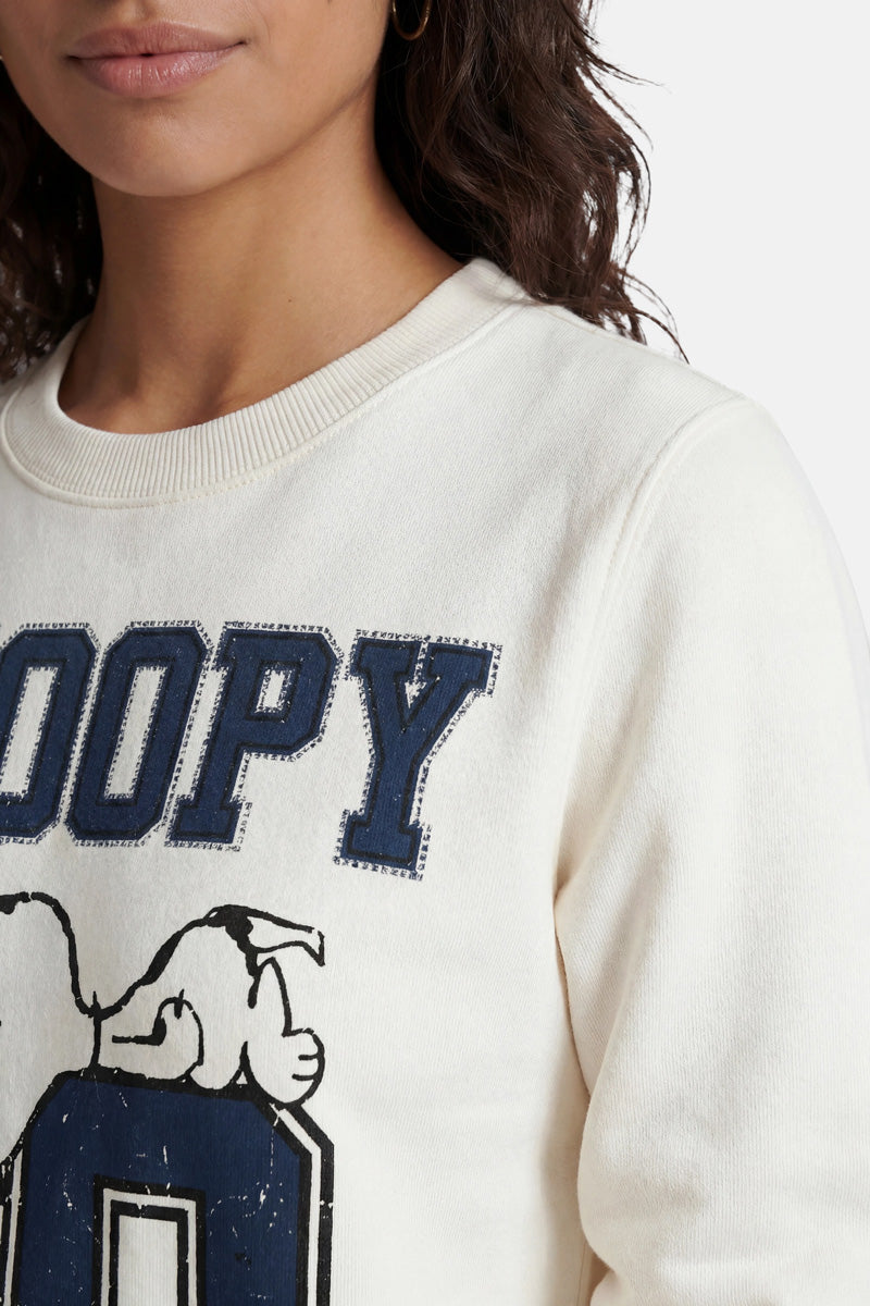 Eine Frau mit welligem braunem Haar trägt einen Snoopy-Sweater aus Baumwolle aus der „Princess Goes Hollywood“-Limited-Edition mit blauem „SNOOPY“-Schriftzug im Used-Look und einem auf dem „O“ liegenden Snoopy vor einem schlichten weißen Hintergrund.