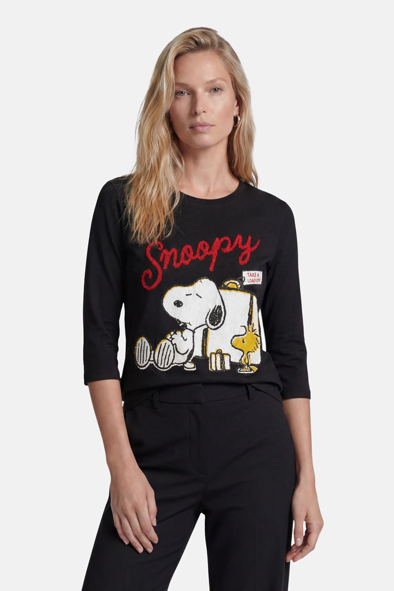 Eine Frau mit langen blonden Haaren trägt ein Langarmshirt aus Baumwolle der Limited Edition „Princess Goes Hollywood“ mit großen Cartoon-Motiven von Snoopy und Woodstock, über denen in roter Schreibschrift „Snoopy“ steht; sie blickt nach vorne und hat eine Hand in der Tasche.