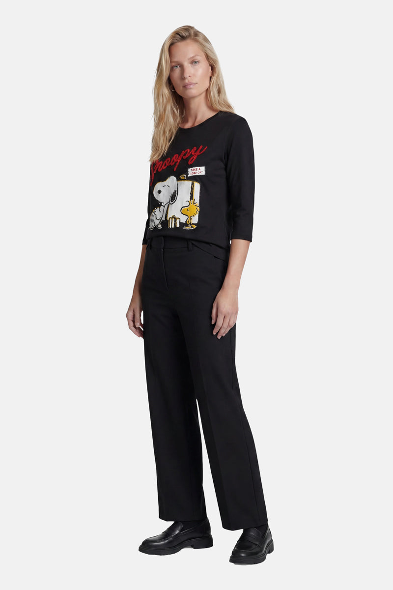 Eine Frau mit langen blonden Haaren trägt ein schwarzes Snoopy-Shirt aus der „Princess Goes Hollywood“-Limited-Edition mit Snoopy und Woodstock darauf, dazu eine schwarze Hose mit weitem Bein und Schuhe. Sie steht entspannt vor einem schlichten hellen Hintergrund und blickt nach vorne.