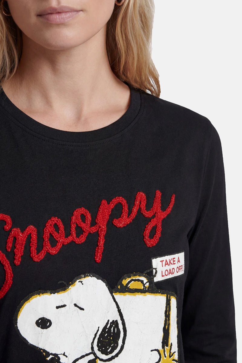 Eine Frau mit heller Haut und langen blonden Haaren trägt ein schwarzes „Princess Goes Hollywood Limited Edition“-Snoopy-Shirt aus Baumwolle mit rotem, strukturiertem „Snoopy“-Schriftzug, einer großen Grafik, auf der Snoopy schläft, und einem weißen Aufnäher mit der auffälligen roten Aufschrift „TAKE A LOAD OFF!“.