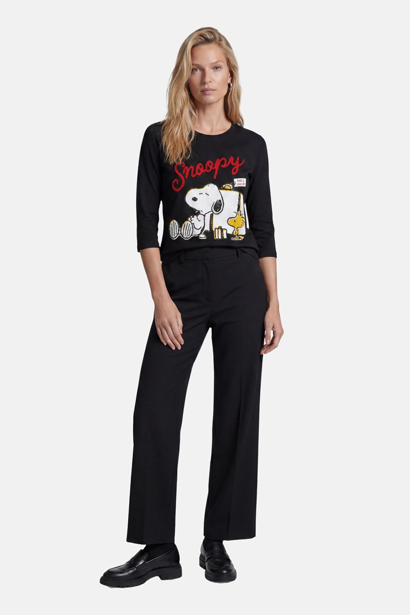 Eine Frau mit langen blonden Haaren trägt ein schwarzes „Princess Goes Hollywood Limited Edition“-Snoopy-Shirt mit Snoopy- und Woodstock-Motiven sowie dem Schriftzug „Snoopy“ in Rot, dazu eine schwarze Hose mit weitem Bein und Schuhe, vor einem weißen Hintergrund.