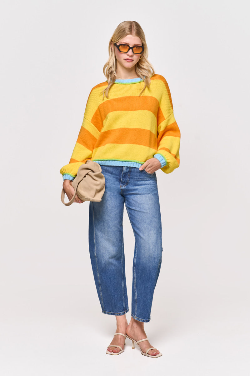 Eine Frau steht vor einem schlichten Hintergrund und trägt einen gelb-orange gestreiften Princess Goes Hollywood-Pullover mit Baumwollmischung und Rundhalsausschnitt, blaue Jeans mit Bündchen, beigefarbene Sandalen, eine Sonnenbrille und hält eine beigefarbene Clutch in der linken Hand.