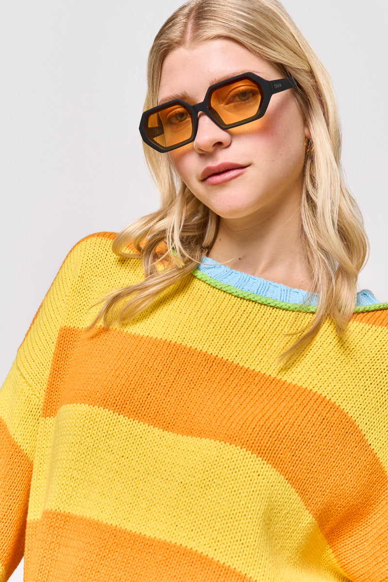 Eine Frau mit langen blonden Haaren trägt eine kantige bernsteinfarbene Sonnenbrille und einen gelb-orange gestreiften Rundhalsausschnitt-Pullover von Princess Goes Hollywood mit blauem Kragen und lächelt leicht in die Kamera vor einem schlichten hellen Hintergrund.