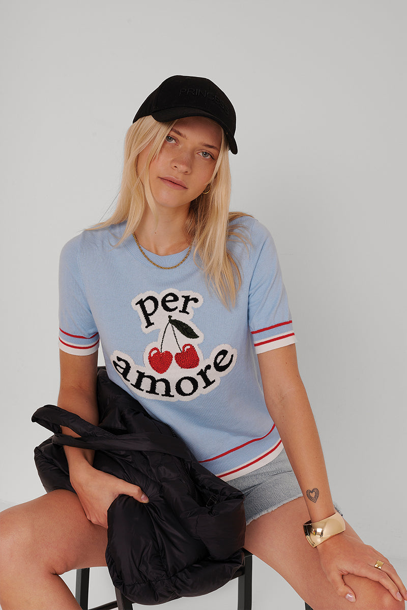 Eine blonde Frau mit schwarzer Mütze, hellblauem Princess Goes Hollywood Per Amore Pullover mit Kirschgrafik, grauen Shorts und Goldschmuck sitzt auf einem Hocker vor einem schlichten Hintergrund. Sie trägt eine schwarze Jacke und sieht entspannt und neutral aus.