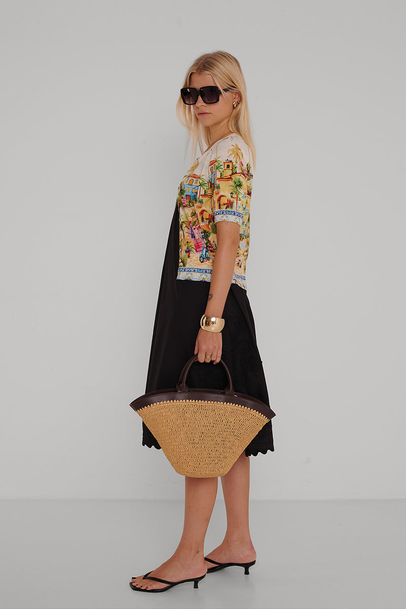 Eine Frau mit langen blonden Haaren und großer Sonnenbrille trägt ein kurzärmeliges Oberteil mit Positano-Print von Princess Goes Hollywood über einem schwarzen Kleid, schwarze hochhackige Sandalen, Goldschmuck und eine große Tragetasche aus Stroh mit dunklen Henkeln.
