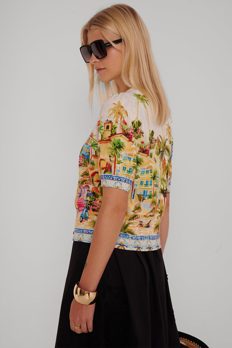 Eine Frau mit glattem blondem Haar trägt eine große schwarze Sonnenbrille, ein kurzärmeliges Oberteil mit Positano-Print von Princess Goes Hollywood, einen schwarzen Rock und ein klobiges Goldarmband. Sie steht vor einem schlichten hellen Hintergrund und schaut über ihre Schulter.