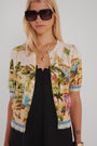 Eine Frau mit glattem blondem Haar trägt eine übergroße dunkle Sonnenbrille, eine goldene Halskette und eine bunte Princess Goes Hollywood Positano-Print Strickjacke mit Ballonärmeln über einem schwarzen Kleid. Sie steht vor einem schlichten hellgrauen Hintergrund.