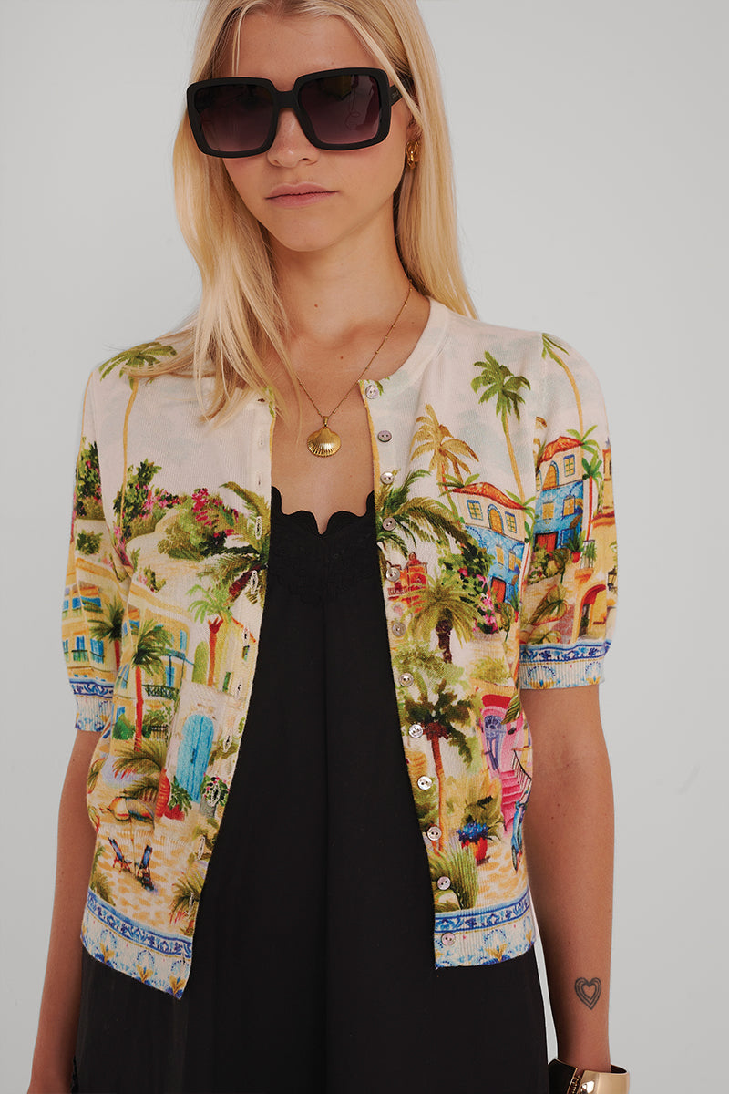 Eine Frau mit langen blonden Haaren trägt eine große schwarze Sonnenbrille, eine goldene Halskette und eine Princess Goes Hollywood-Strickjacke mit der Stickerei eines tropischen Dorfes über einem schwarzen Midikleid. Sie hat einen neutralen Gesichtsausdruck und ein kleines Herz-Tattoo auf ihrem linken Unterarm.