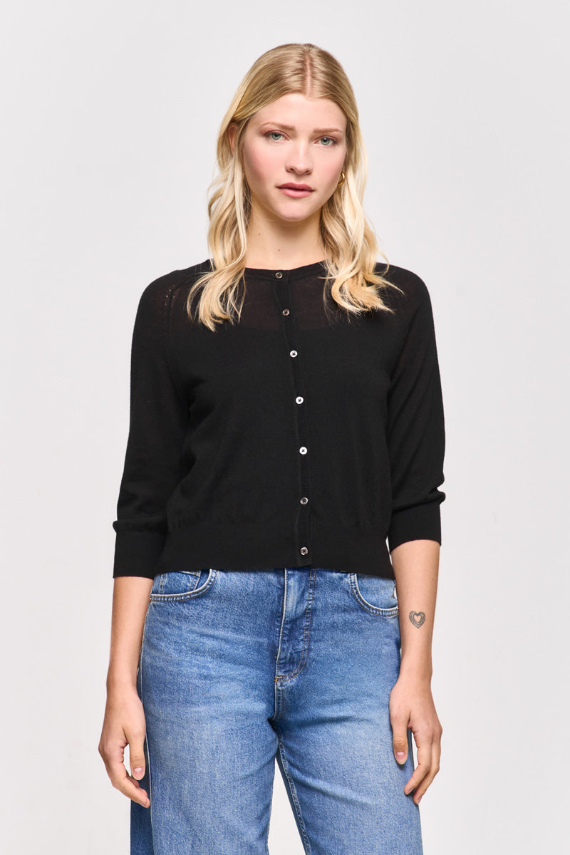 Eine Frau mit langen blonden Haaren trägt eine schwarze Princess Goes Hollywood Strickjacke aus Wolle-Seide-Mix mit Dreiviertelärmeln und blaue Jeans. Sie steht mit dem Gesicht nach vorne vor einem schlichten weißen Hintergrund und zeigt ein kleines Herz-Tattoo auf ihrem linken Unterarm.