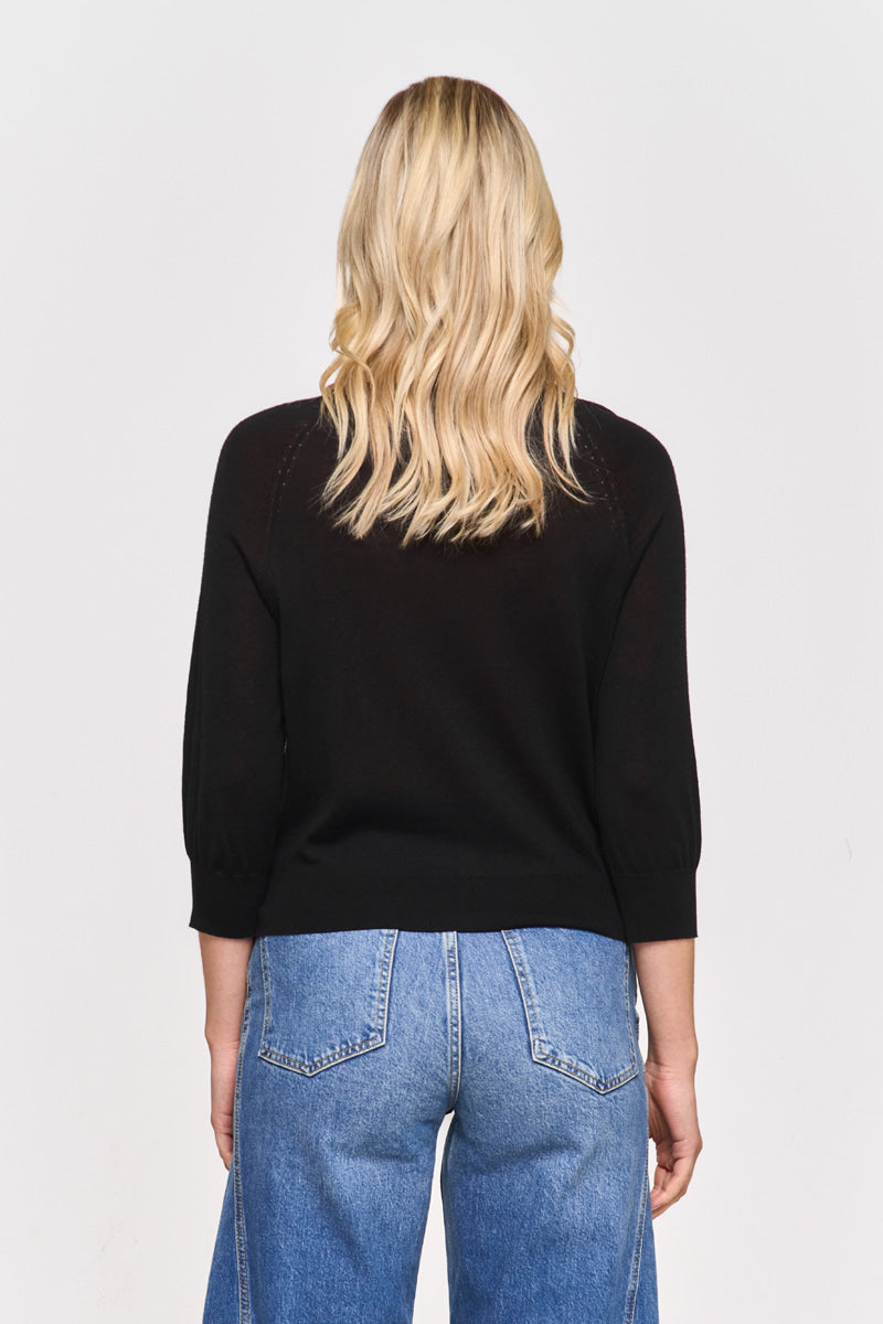 Eine Person mit langem, gewelltem blondem Haar blickt von der Kamera weg und trägt ein schwarzes Princess Goes Hollywood-Top mit Rundhalsausschnitt und Dreiviertelärmeln sowie eine Jeans mit hoher Taille vor einem schlichten hellen Hintergrund.
