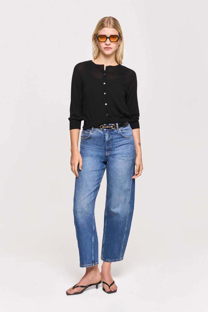 Eine blonde Frau trägt eine braune Sonnenbrille, eine schwarze Princess Goes Hollywood Strickjacke aus Wolle-Seide-Mix, blaue High-Waisted Jeans mit schwarzem Gürtel und schwarze Flip-Flops. Sie steht mit dem Gesicht nach vorne vor einem schlichten weißen Hintergrund.
