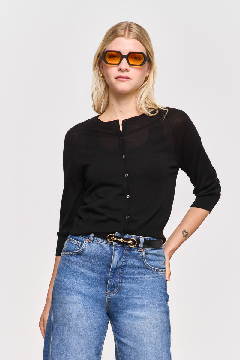 Eine Frau mit blondem Haar trägt eine orangefarbene Sonnenbrille und eine Princess Goes Hollywood-Strickjacke mit Dreiviertelärmeln, schwarzen Knöpfen, Bluejeans mit hoher Taille und einem schwarzen Doppelringgürtel vor einem schlichten hellen Hintergrund.