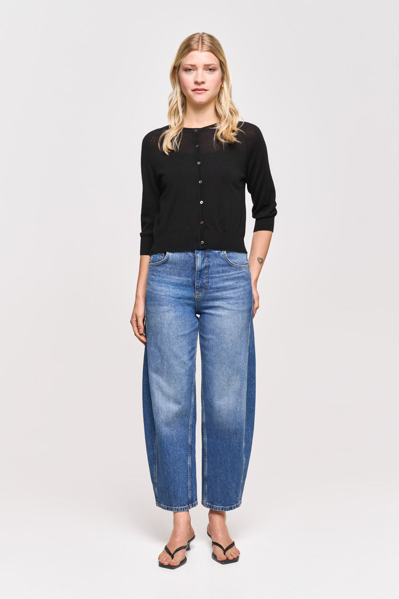 Eine Frau mit langen blonden Haaren blickt nach vorne und trägt eine schwarze Princess Goes Hollywood-Strickjacke aus einem Woll-Seiden-Mix, blaue Jeans mit weitem Bein und schwarze Sandalen. Sie hat eine kleine Armtätowierung, die Arme liegen entspannt an ihren Seiten, vor einem schlichten weißen Hintergrund.