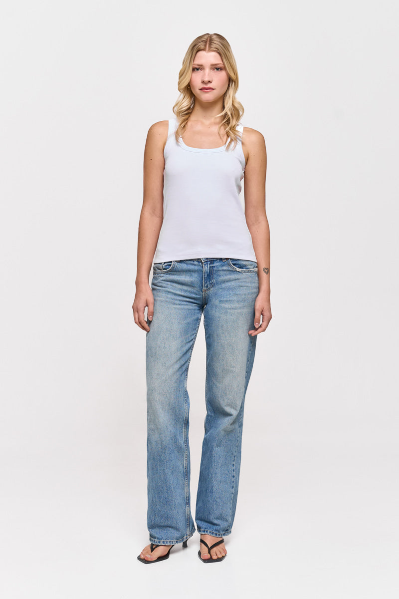 Eine Frau mit gewelltem blondem Haar steht mit dem Gesicht nach vorne vor einem weißen Hintergrund und trägt ein weißes Princess Goes Hollywood Basic Top aus Baumwolle mit Rundhalsausschnitt, hellblaue Jeans und schwarze Flip-Flops. Ihre Arme sind entspannt und ihr Gesichtsausdruck ist neutral.