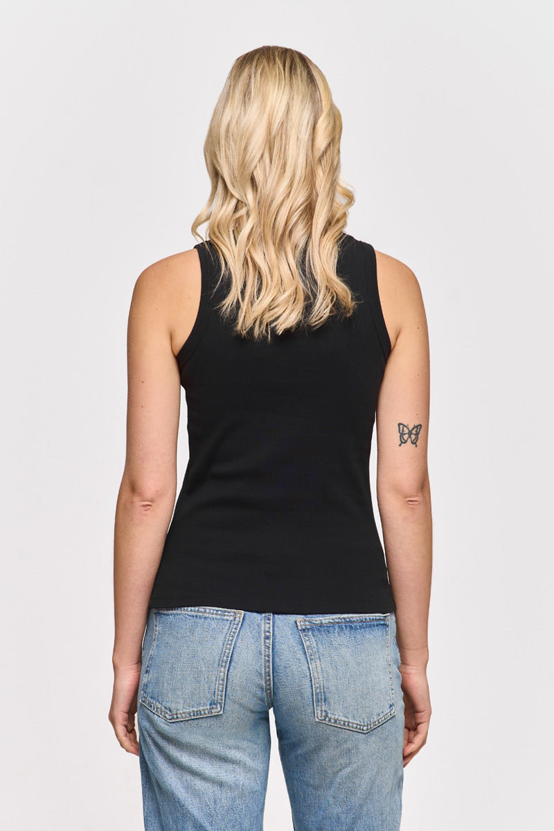 Eine Frau mit gewelltem, schulterlangem blondem Haar ist von hinten zu sehen. Sie trägt ein schwarzes Princess Goes Hollywood Basic Top mit Rundhalsausschnitt und hellblaue Jeans. Ein Schmetterlingstattoo ziert ihren linken Oberarm. Der Hintergrund ist einfarbig weiß.